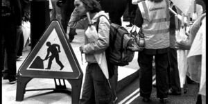 Black and white image of a woman walking past a protest.