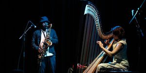 A black man stands playing a saxophone, and next to him a woman plays a harp