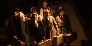 An image of a group of actors on stage holding briefcases, dressed in period clothing.