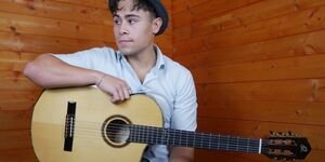 A young man wearing a grey hat and white shirt sits with a guitar on his lap