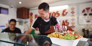 A phot of a glass counter on top of which sits a burrito. Two pepple can be seen in the background dressed in black