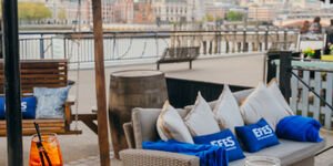 The outside of the bar with a beige sofa with cusions, and drinks on a table. In the background is a view of the river Thames