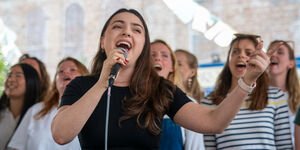A woman in a black tshirt stands holding a microphone singing. Behind her are a group of women standing and singing