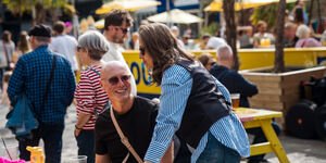A man sits at a picnic bench in the sunshine with a drink, whilst a woman stands next to him