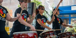 4 people in black t-shirts stand in font of large red drums holding drumsticks