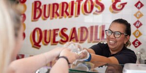 A woman stands behind a food counter passing a box of food to a person in front