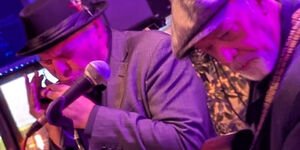 A man plays a harmonica and another plays a guitar. Both are standing in front of a microphone