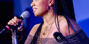 A woman with long black hair stands at a microphone, holding it