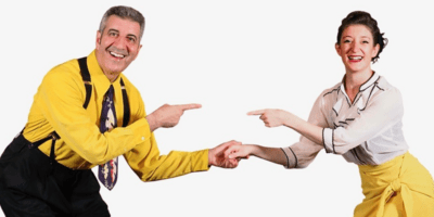 A man and a woman dressed in black, yellow and white hold hands and point at eachother, smiling